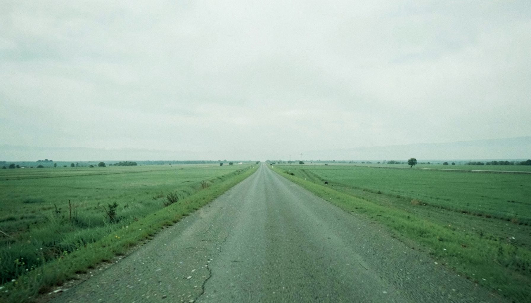 A wide, muted green landscape suggesting a long highway corridor stretching into the distance under a soft overcast sky, calm and open