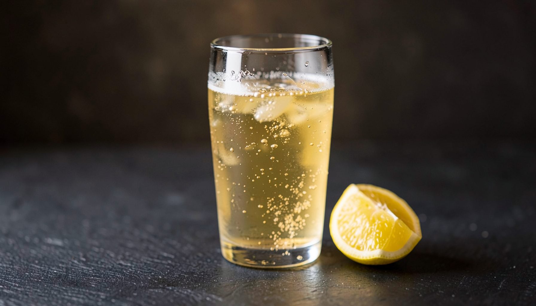 A glass of pale golden craft-brewed hard seltzer on a dark slate surface, with subtle bubbles rising and a wedge of lemon.
