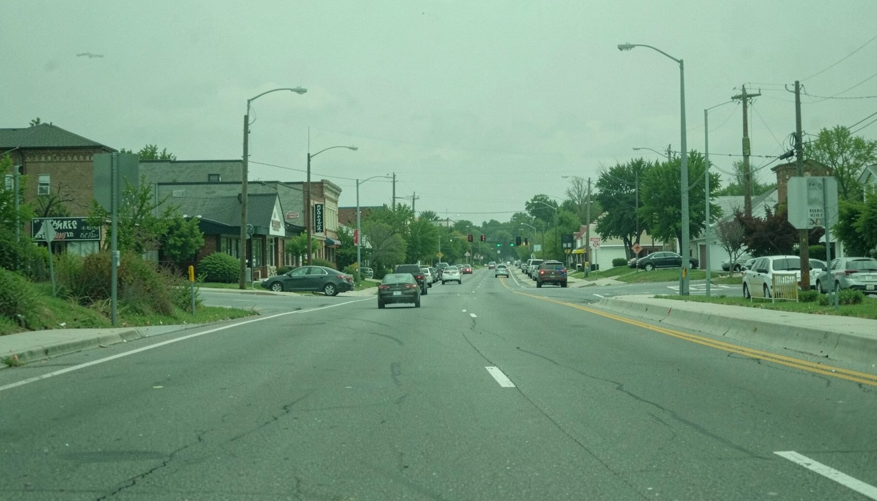 A muted green highway landscape with rows of small storefronts and parked cars along a suburban road.