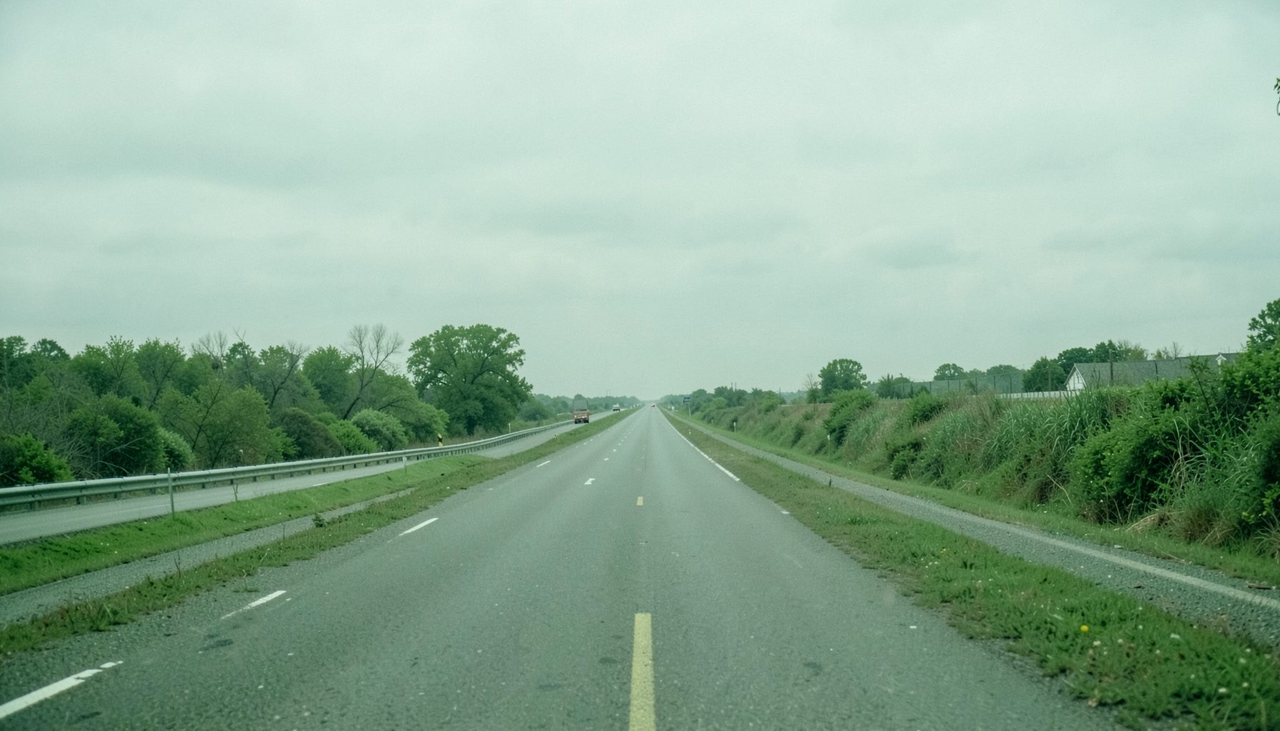 A wide, muted green landscape suggesting a long highway corridor stretching into the distance under a soft overcast sky.